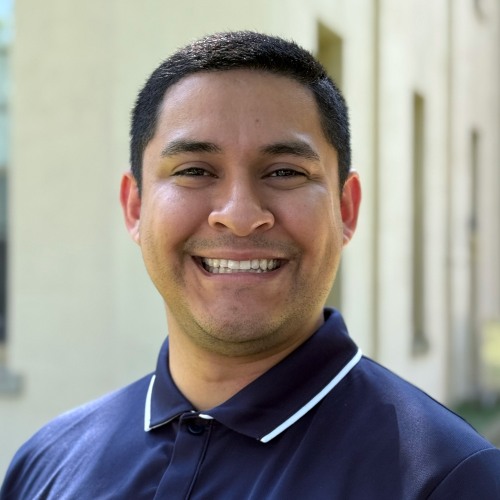 Headshot of Joshua Santos wearing a navy blue Brown University polo shirt, standing outside of a building