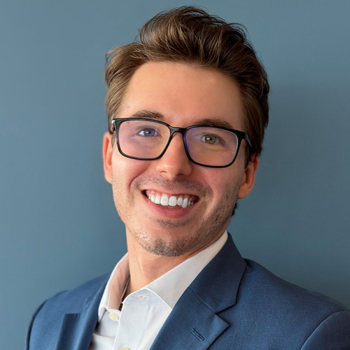 Sam Sumeracki standing in front of a blue wall, wearing a suit and smiling