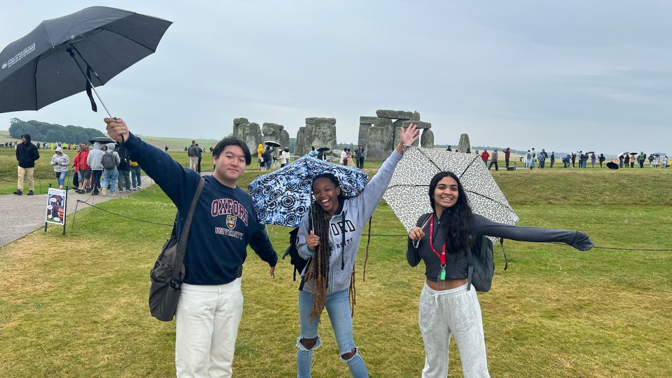 Students standing in front of Stonehenge on a rainy day with umbrellas